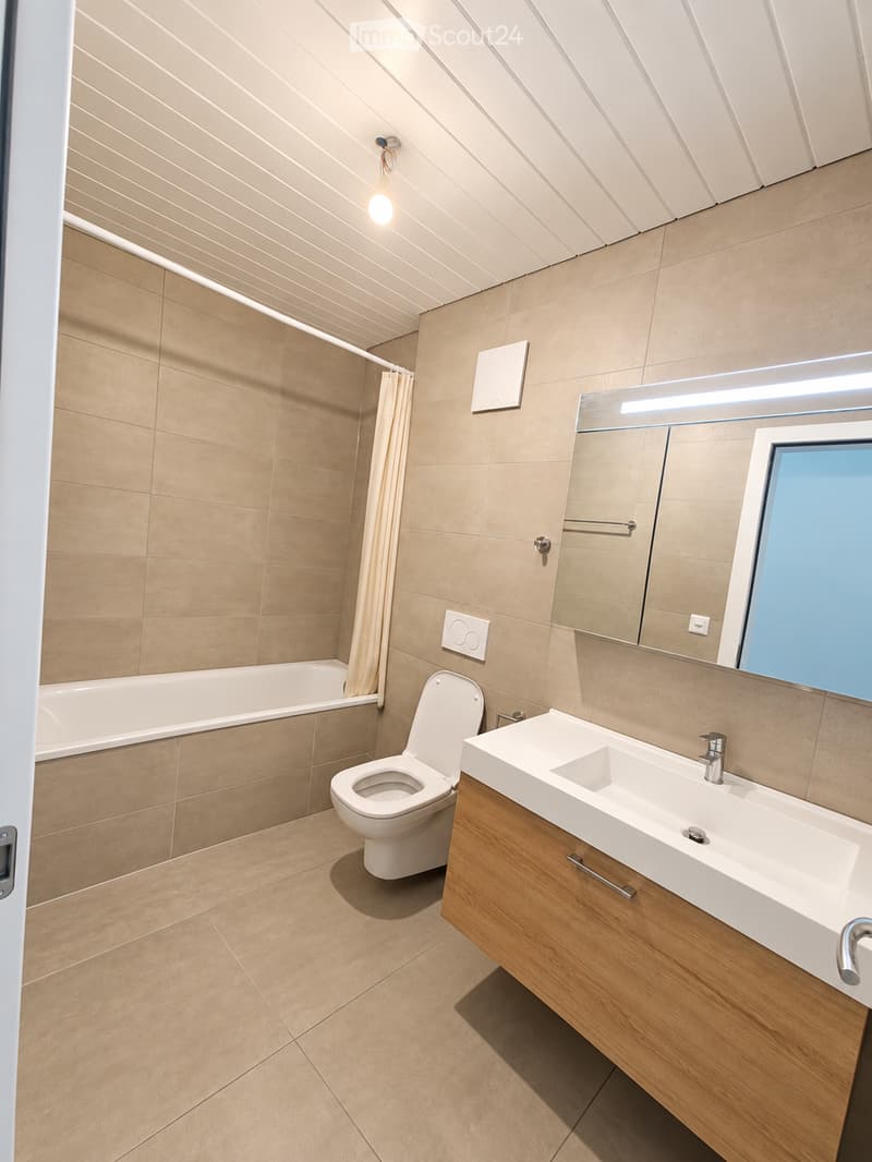 Modern bathroom with beige tiles, a white toilet, a wooden sink cabinet, a bathtub, and a large mirror with built-in lighting.