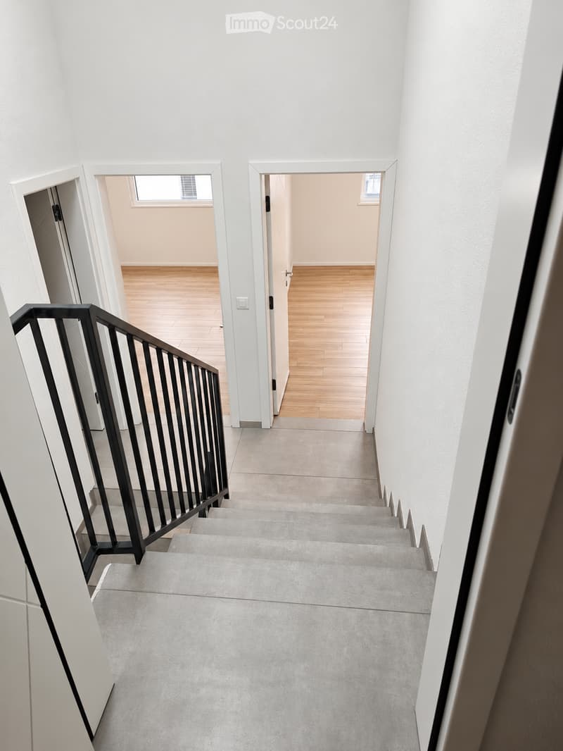 Modern hallway with a staircase leading upwards, featuring grey carpeted stairs, a black metal railing, and white walls. Two open doorways lead to rooms with wooden flooring and small windows.