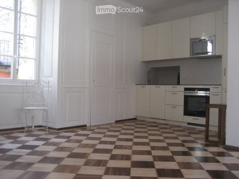Modern kitchen with white cabinets, stainless steel appliances, and a checkered floor. Features a small wooden table and a white metal chair.
