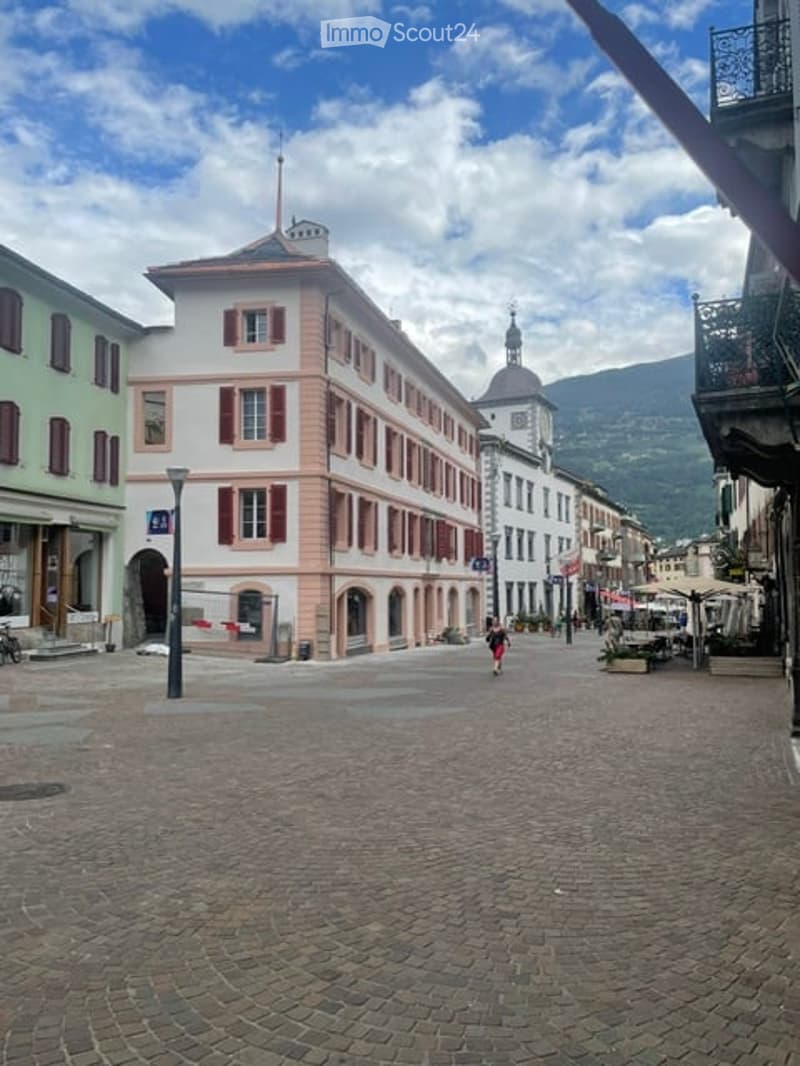 A charming European town square with a multi-story building featuring red shutters and a dome-shaped roof, surrounded by other historic buildings and a scenic mountain backdrop.