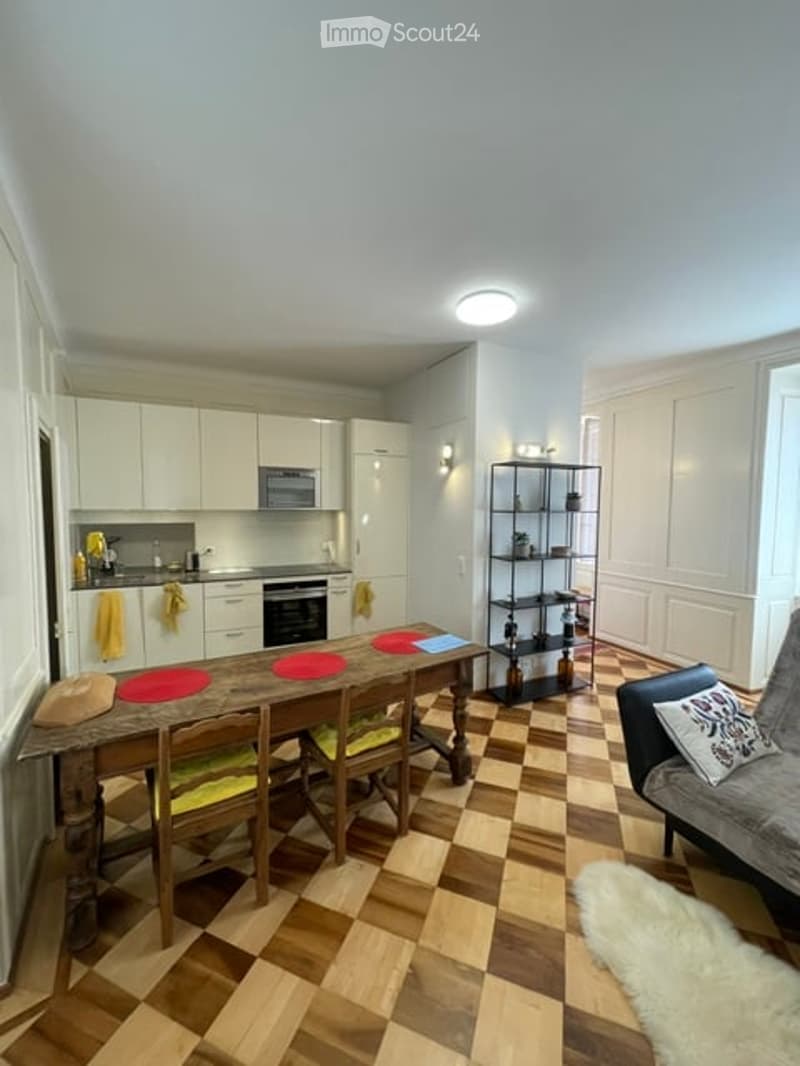 A modern, open-plan kitchen and living area with white cabinets, a wooden dining table, and a comfortable sofa. The space features a checkered floor and a sleek, contemporary design.