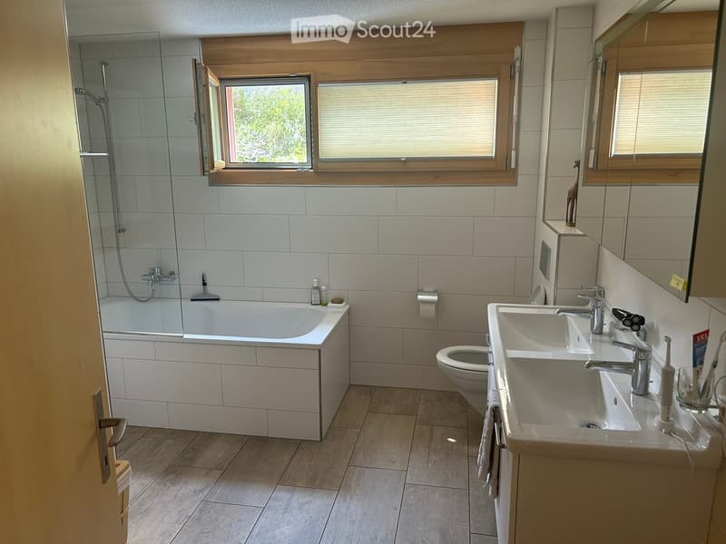 Modern bathroom with a bathtub, double sink, toilet, and large windows. Includes a mirror, toothbrush holder, and towel.