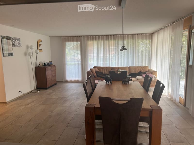 Bright living and dining area with large windows and a balcony view, featuring a sofa, dining table with chairs, and a wooden cabinet.