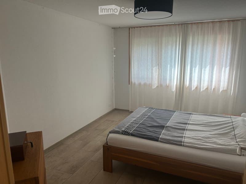 Minimalist bedroom with a single bed, nightstand, and sheer curtains on a window. The room features light-colored walls and wooden flooring.