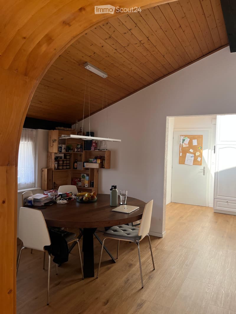 A modern, well-lit room with wooden floors and a slanted wooden ceiling. Features a round wooden dining table with four chairs, a laptop, a water bottle, a cup, and a bowl of fruit. Shelves hold various items, and a door leads to another room.