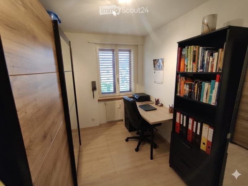 A well-lit study or office space with wooden flooring, a large window with blinds, a desk with a laptop, a chair, a bookshelf filled with books and binders, and a printer on the desk.