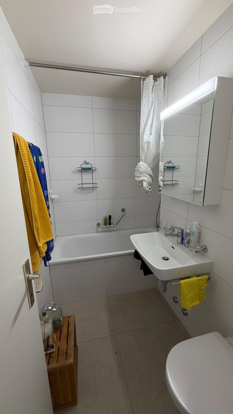 Modern bathroom with white tiles, bathtub, sink, toilet, mirror, and shelves. Towels and toiletries are visible.