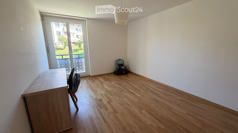 A small, unfurnished room with wooden flooring, a desk, a chair, and a backpack near the window.