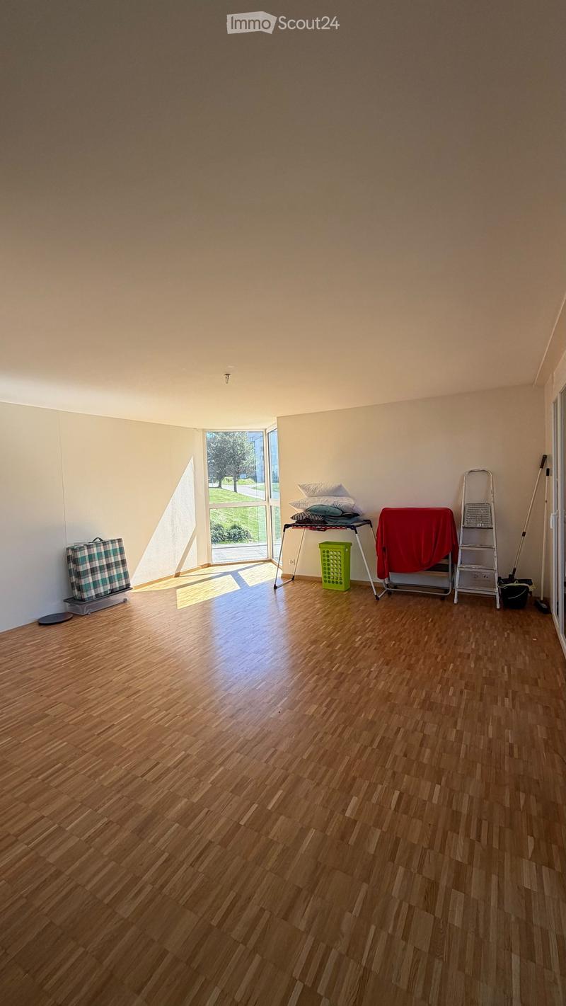 Empty room with wooden flooring, large window, and some storage items including a stool, ladder, and suitcase.