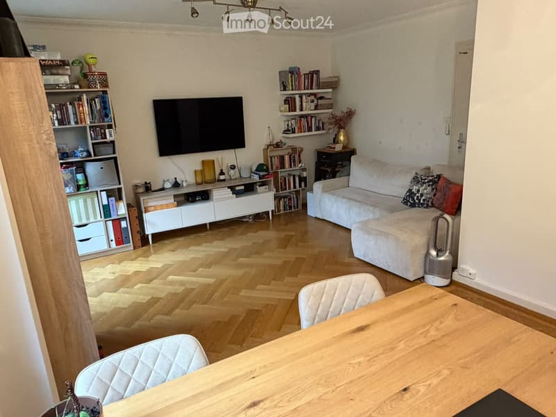 Modern living room with light wooden flooring, a white sofa, a wooden dining table with two chairs, a TV mounted on the wall, and several bookshelves filled with books and decorative items.