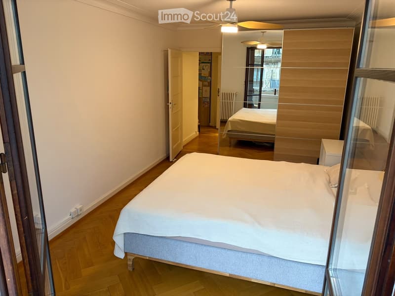 Spacious bedroom with wooden flooring, large bed with white bedding, nightstand, and open door leading to another room.
