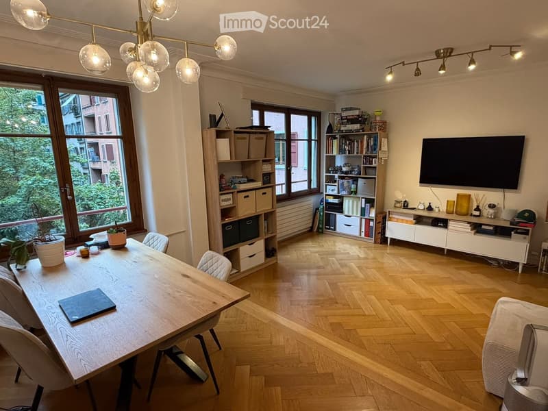 Bright, modern room with wooden flooring, large windows, a dining table with chairs, a laptop, a bookshelf, a TV, and decorative items.