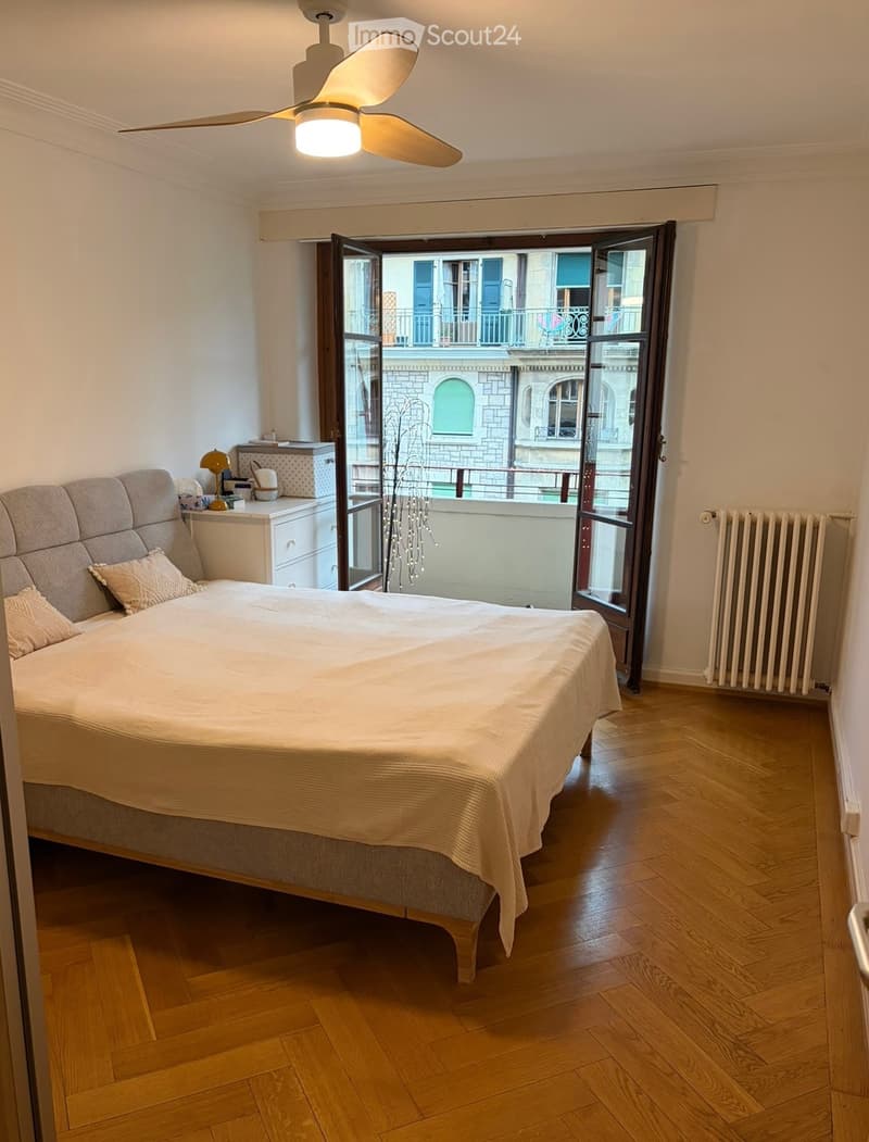 Bedroom with wooden flooring, large bed with pillows and blanket, nightstand with lamp, radiator, and open sliding doors leading to a balcony.