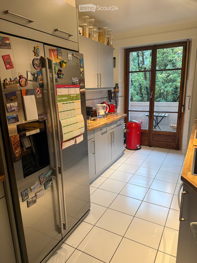 Modern kitchen with white tiled floor, grey cabinets, stainless steel refrigerator adorned with magnets and photos, a wooden countertop with a blender, and a red trash bin near the door leading to a balcony.