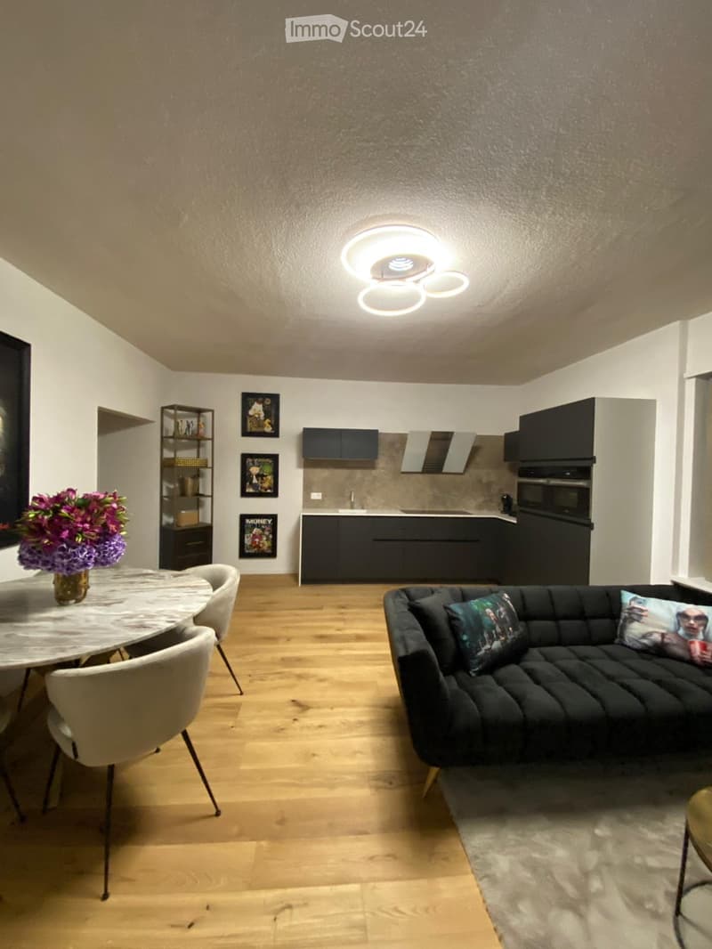 Modern open-plan living and dining area with a dark grey sofa, a round marble dining table with four chairs, a vase with purple flowers, a kitchen area with dark cabinets and stainless steel appliances, and a sleek ceiling light fixture.