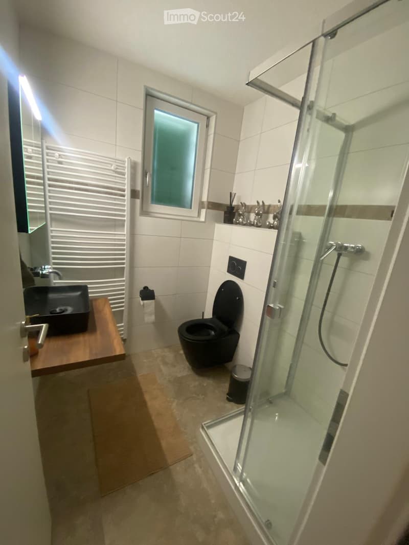 Modern bathroom with white tiles, a black sink, toilet, and glass-enclosed shower. Features a wooden vanity, towel rack, and a window with blinds.