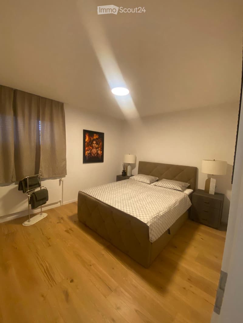 Cozy bedroom with wooden flooring, a large bed with patterned bedding, two nightstands with lamps, a picture frame on the wall, and a small chair near the window.