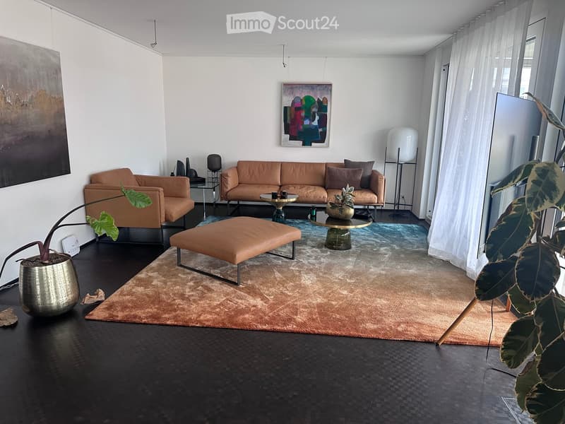 Modern living room with a tan leather sofa, ottoman, coffee table, side tables, floor lamp, and potted plants. The room features large windows with sheer curtains and a dark wooden floor.