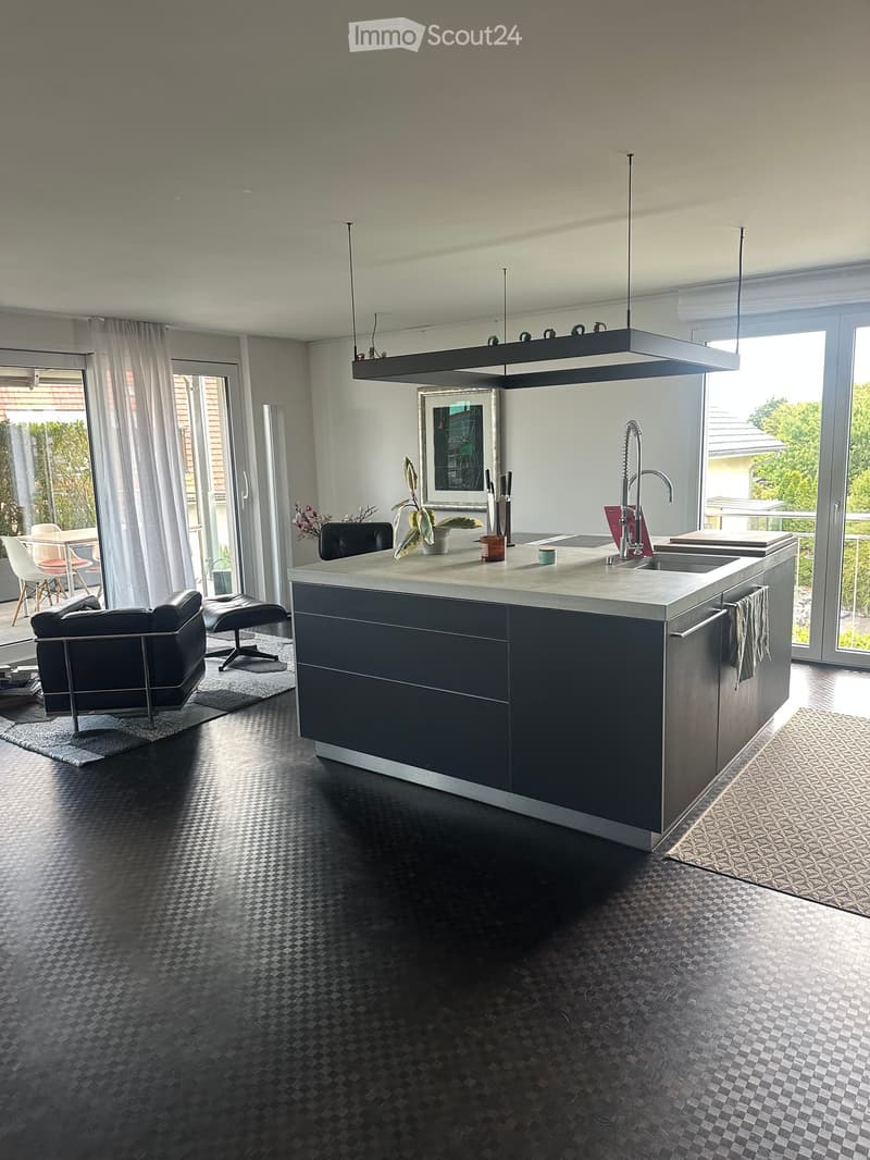 Modern kitchen with a central island, sleek black cabinets, and a dark tiled floor. Features include a stainless steel faucet, a potted plant, and a small seating area with a chair and table.