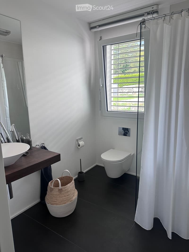 Modern bathroom with white walls, black tiled floor, sink with wooden countertop, toilet, mirror, and wicker basket on floor.