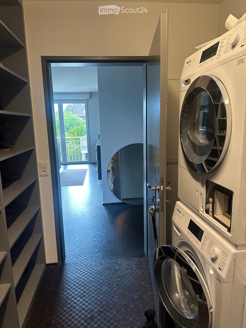 A hallway with a washing machine and dryer on the right side, a door leading to a balcony area with a view of greenery, and a shelf unit on the left side.