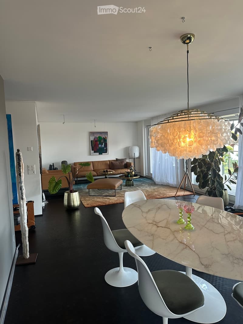 Modern living and dining area with a black floor, white chairs, a marble dining table, a brown sofa, and a large chandelier.