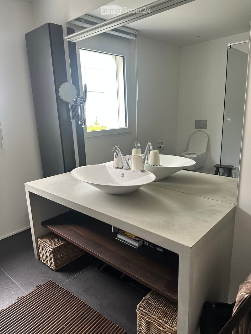 Modern bathroom with a double sink vanity, sleek faucets, a large mirror, and a toilet. The vanity has a light-colored countertop and storage space underneath, with wicker baskets visible.