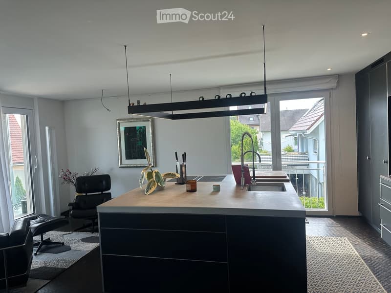 Modern kitchen with a central island, dark cabinets, pendant lighting, and a sleek faucet. Includes a black leather chair and stool, a potted plant, and a framed picture on the wall.