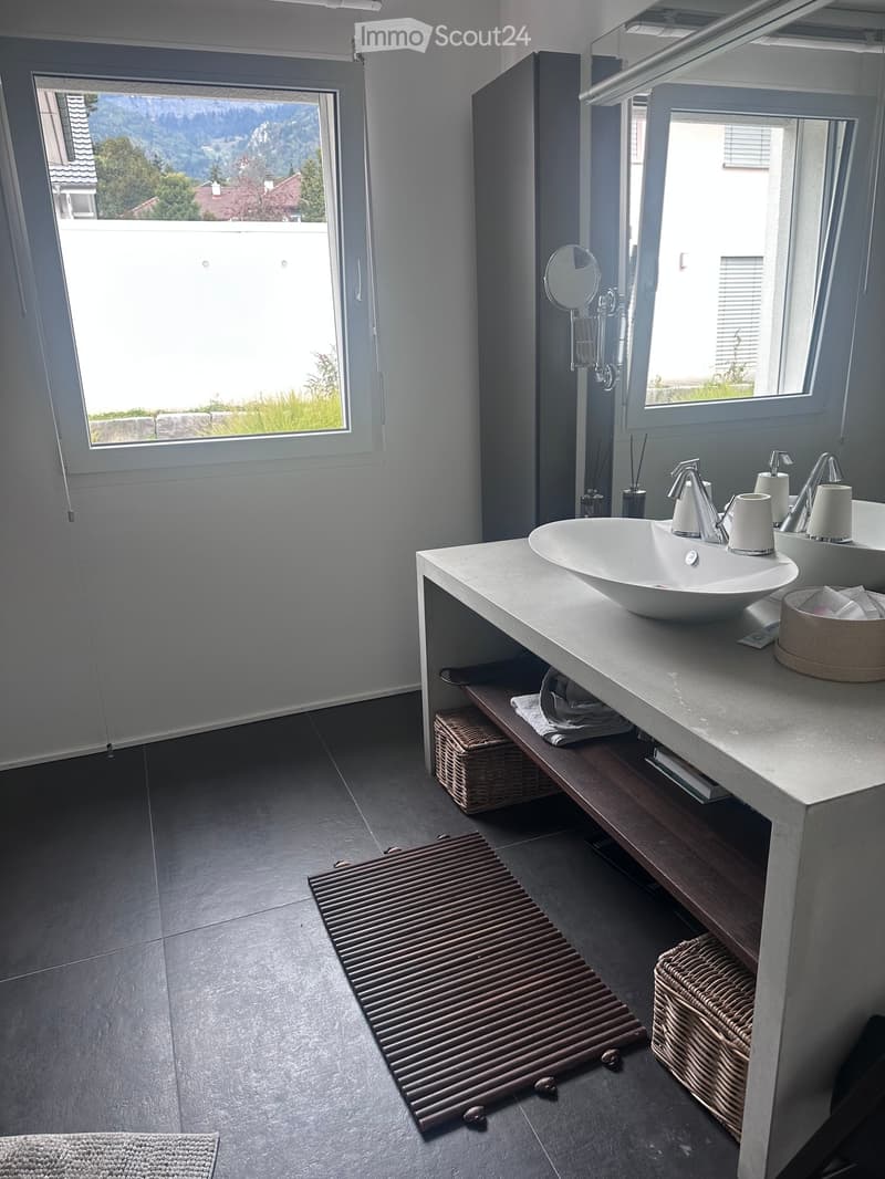 Modern bathroom with double sink vanity, sleek faucets, large windows offering a scenic view, wicker baskets, and a wooden bath mat.