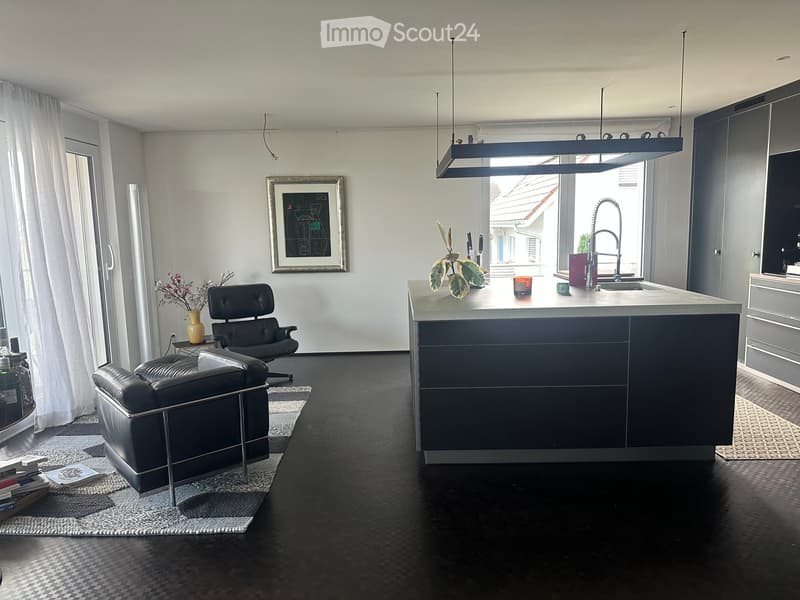 Modern open-plan living and kitchen area with dark wood flooring, a black leather sofa, a kitchen island, and a sleek faucet. The space features contemporary furniture, a framed picture on the wall, and a potted plant on the island.