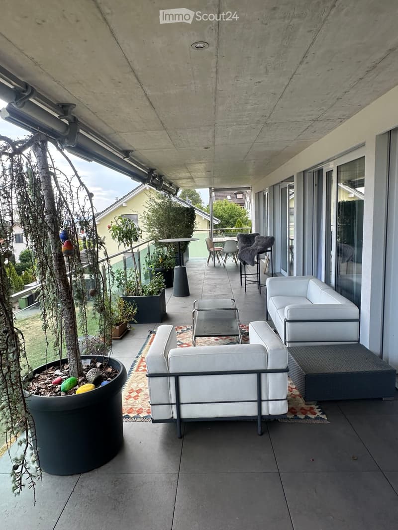 Modern balcony with white sofa, chairs, small table, and decorative rug, surrounded by potted plants and trees.