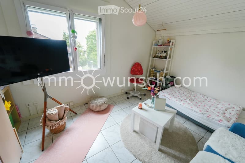 Cozy bedroom with a bed, small table, chair, shelf, and a pink exercise mat. Large windows provide natural light, and a TV is mounted on the wall.