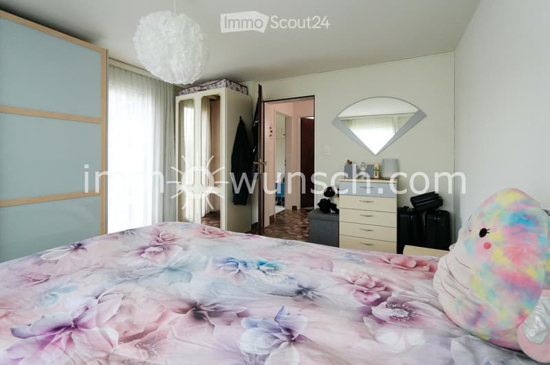 A well-lit bedroom with a large bed covered in a floral duvet, a colorful stuffed toy, a wardrobe, a nightstand with a lamp, and a mirror on the wall.
