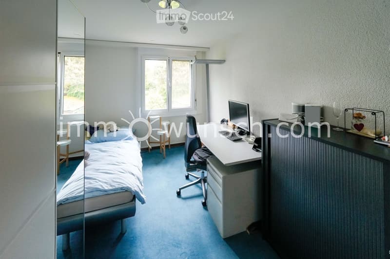 A well-lit bedroom with a bed, desk, and chair. The room features a large mirror, a window with blinds, and a blue carpeted floor.