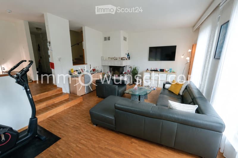 Modern living room with hardwood flooring, grey sectional sofa, glass coffee table, armchair, fitness bike, fireplace, TV, and cabinet.