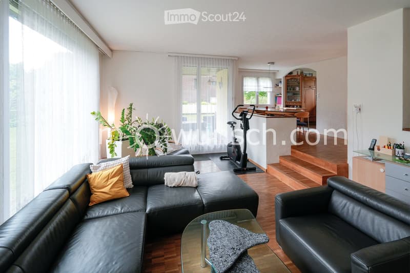Modern living room with a black leather sectional sofa, glass coffee table, exercise bike, and a small kitchen area with cabinets and a counter.