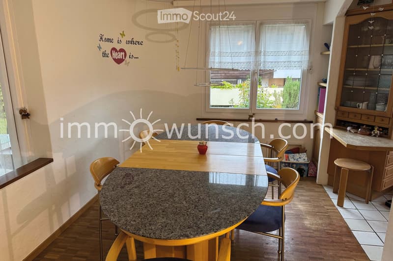 The image shows a well-lit dining area with a round granite table surrounded by six wooden chairs. There is a wooden sideboard with glass-fronted cabinets and a small stool. The room features wooden flooring, large windows with white curtains, and a decor