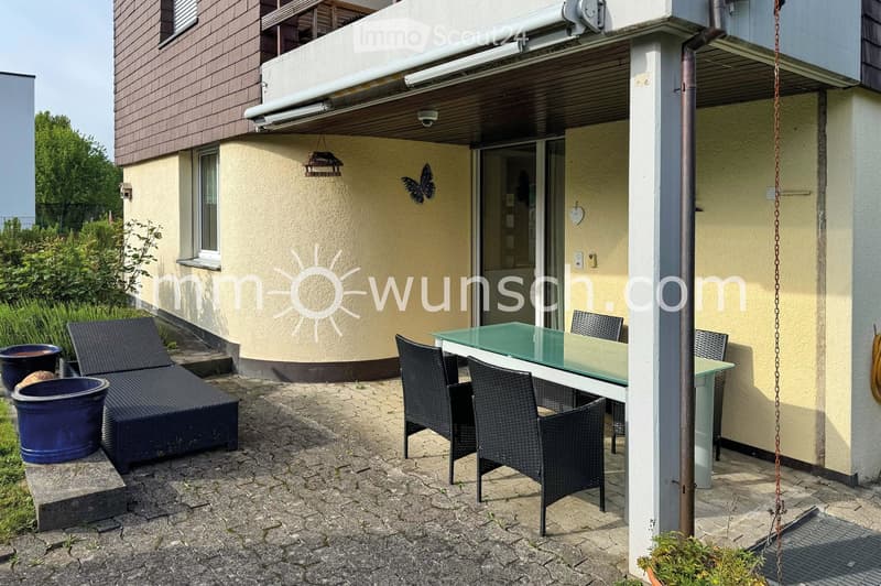 Outdoor patio area with a glass table, wicker chairs, and a dark-colored sofa, surrounded by potted plants and a covered entrance to the building.