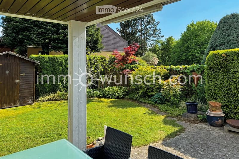 Well-maintained garden with a covered patio area, green lawn, various shrubs and trees, and outdoor furniture including a glass table and wicker chairs.
