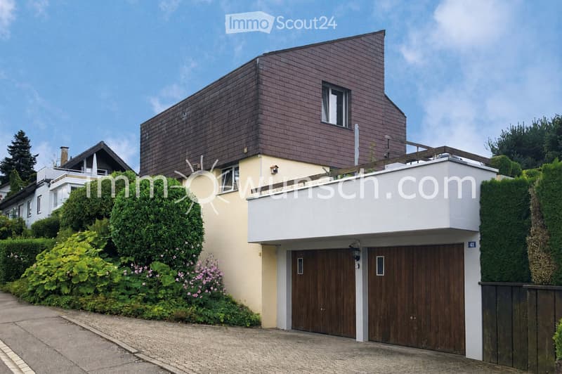 Modern two-story house with a combination of brown and white exterior walls, large wooden garage doors, and a well-maintained garden with flowering shrubs.