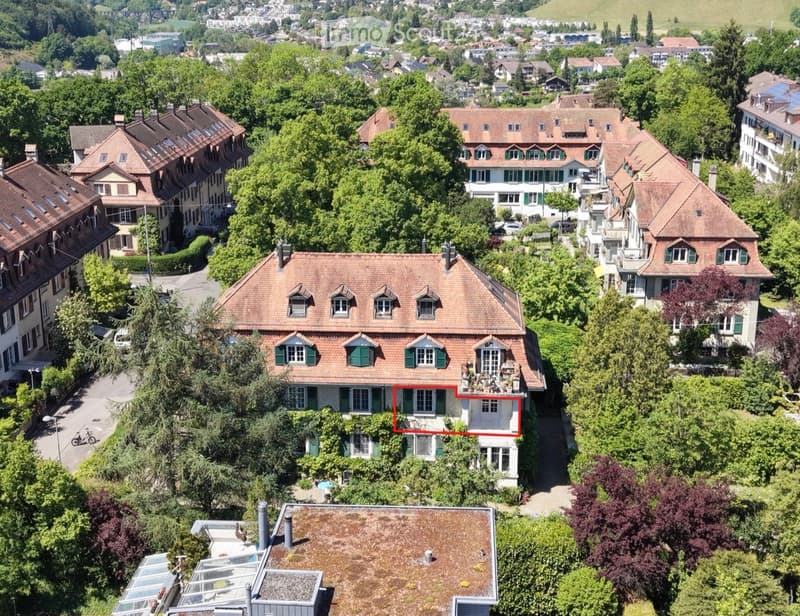 Aerial view of a residential neighborhood with multiple houses, surrounded by lush greenery and trees. The houses have red-tiled roofs and are interspersed with trees and shrubs.