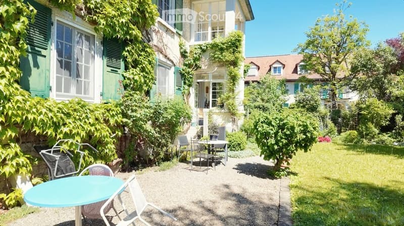 A serene garden area with a small patio seating arrangement, including a turquoise table and several chairs, surrounded by lush greenery and a charming house with ivy-covered walls.