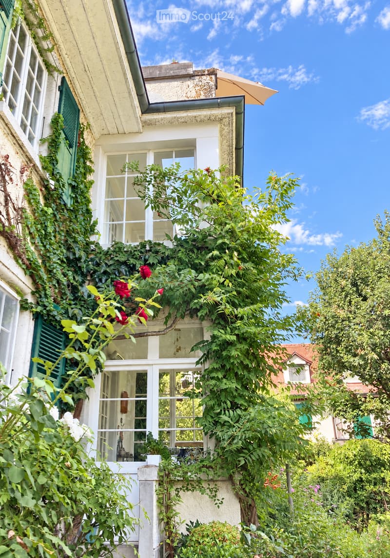 A charming garden view with lush greenery, climbing vines, and a small balcony area with white railings and a window overlooking the garden. The background features a classic building with green shutters and a clear blue sky.