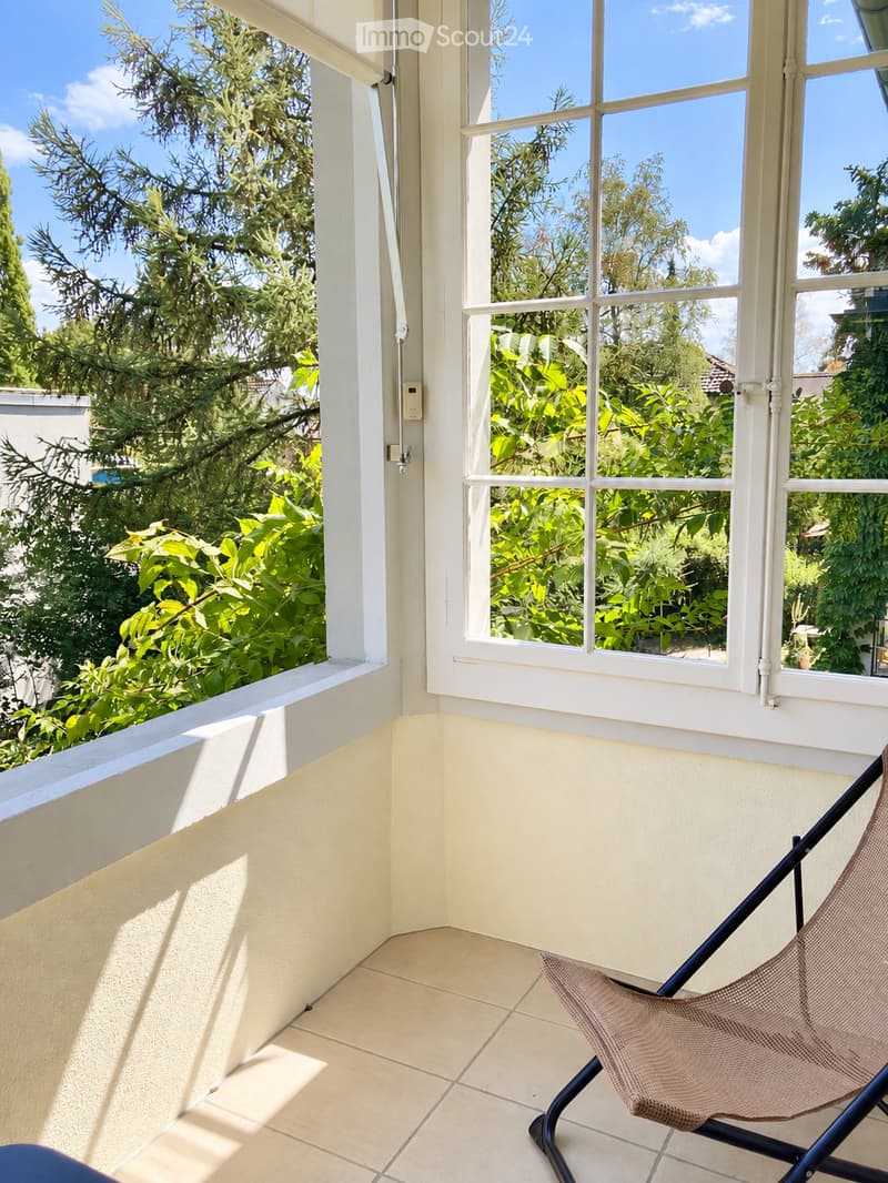 Balcony with tiled floor, white walls, large windows overlooking a garden, and a single folding chair.