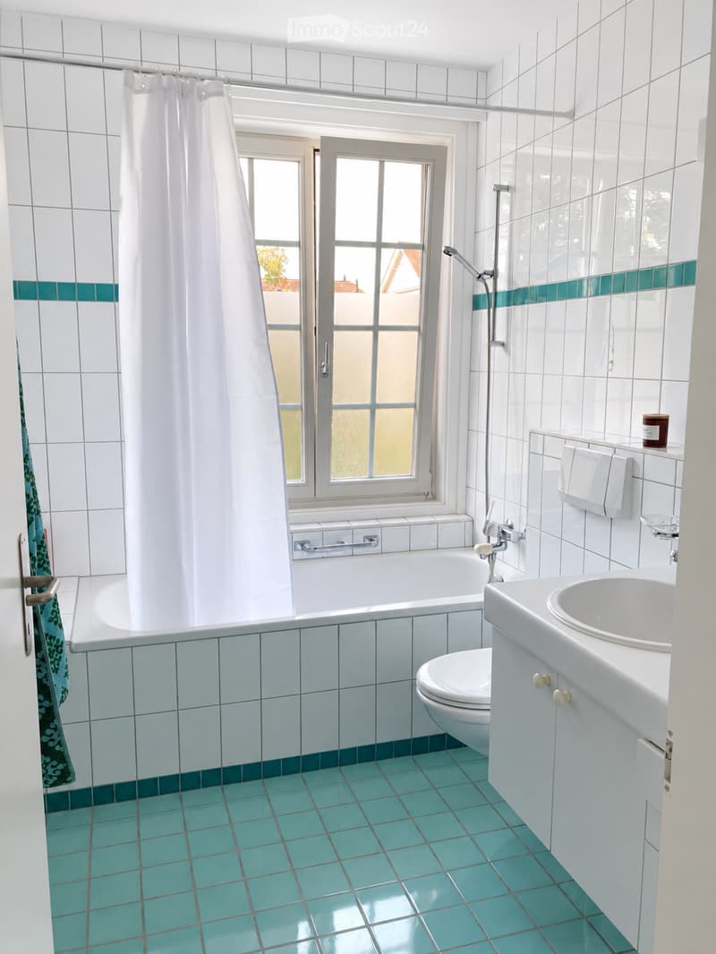 Bright bathroom with white tiled walls, turquoise tiles on the floor, a bathtub, toilet, sink, and a window with a shower curtain.