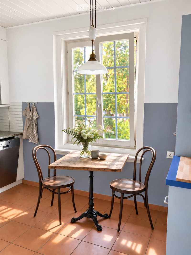 Bright kitchen with a wooden dining table, two chairs, a vase with white flowers, and a hanging pendant light. The space features blue walls, orange tiled flooring, and a view of greenery through a large window.