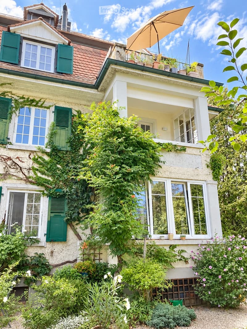 A charming garden view of a two-story house with ivy-covered walls, green shutters, and a balcony with potted plants and an umbrella.