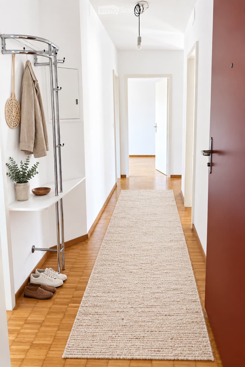 Modern hallway with wooden flooring, white walls, a long beige runner, coat rack with coats, shoes, small shelf with plant and bowl, and a hanging light fixture.