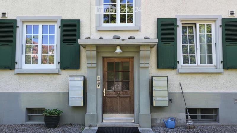Facade of a two-story building with a wooden front door, green shutters, and multiple windows. There are mailboxes on either side of the entrance, a potted plant, a broom, and a watering can visible near the door.
