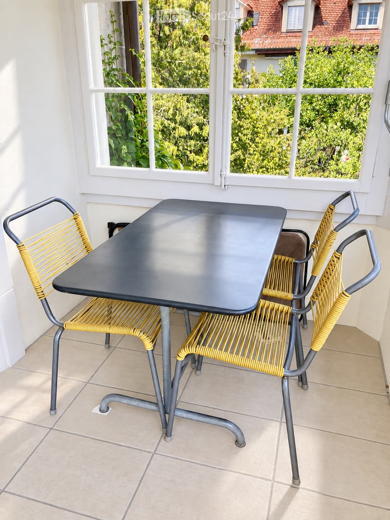 Balcony with a black table and two yellow chairs, large windows with a view of greenery.